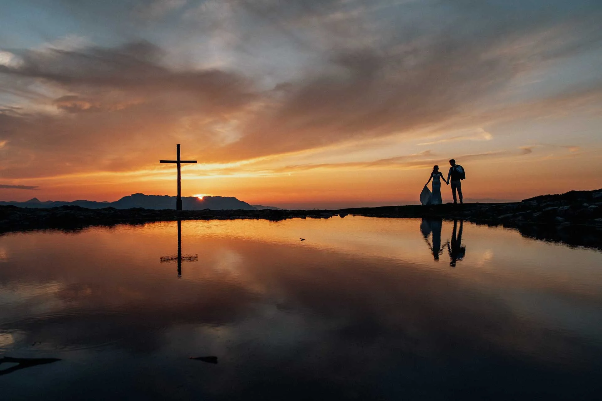 Couple backlit at a mountain lake with a summit cross